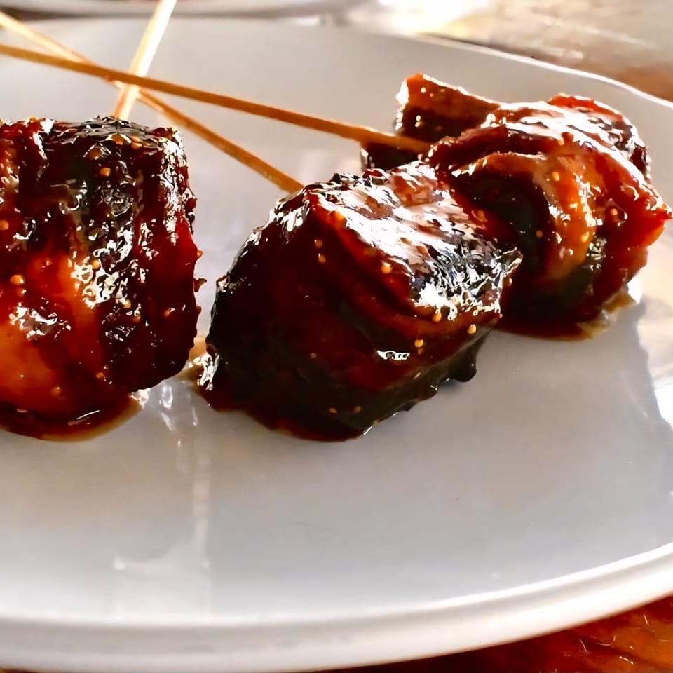 Three skewers of American street food, possibly pork belly, coated in a dark, glossy sauce on a white plate.