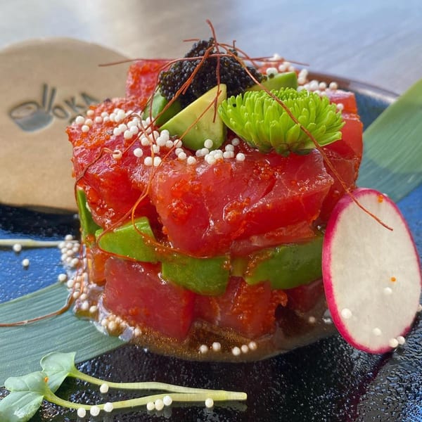 Tuna poke with avocado, black caviar, white pearls, and a radish slice on a dark plate.