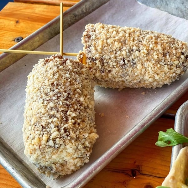 Two American corn dogs, coated in crushed nuts, served on a metal tray with parchment paper.