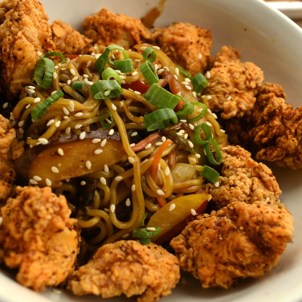 A close-up of Lilo's Streetfood's fried chicken and noodles with sesame seeds and green onions.