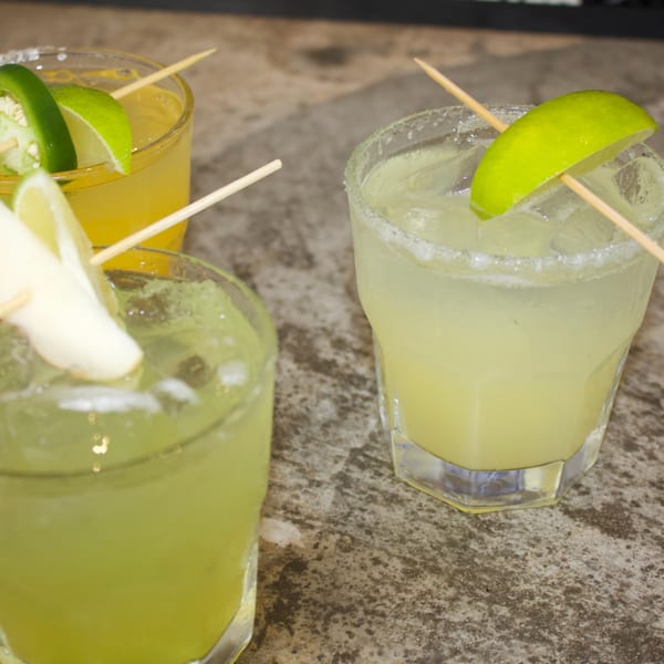 Three vibrant cocktails, two green and one yellow, garnished with lime, jalapeño, and ginger.