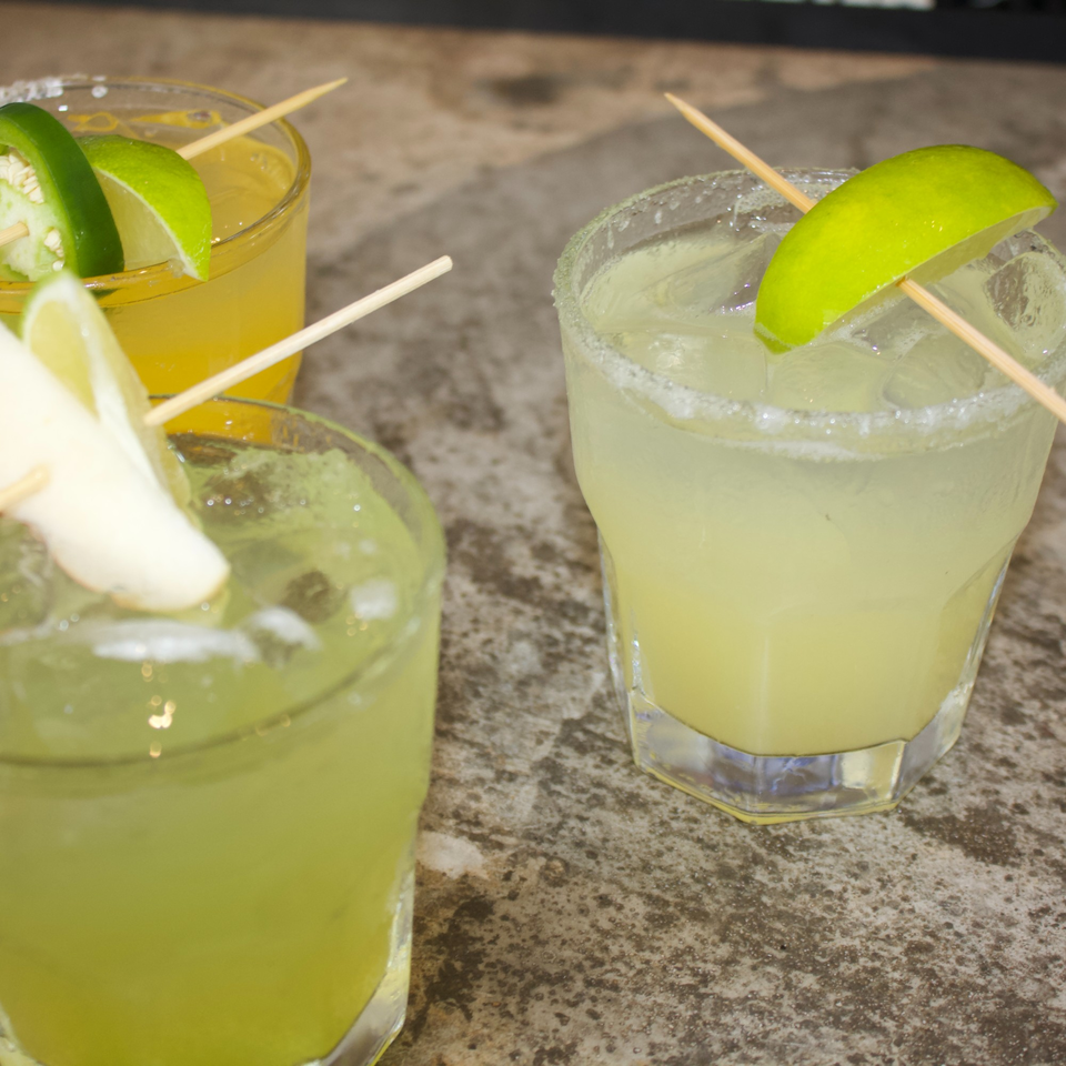 Three vibrant cocktails, two green and one yellow, garnished with lime, jalapeño, and ginger.