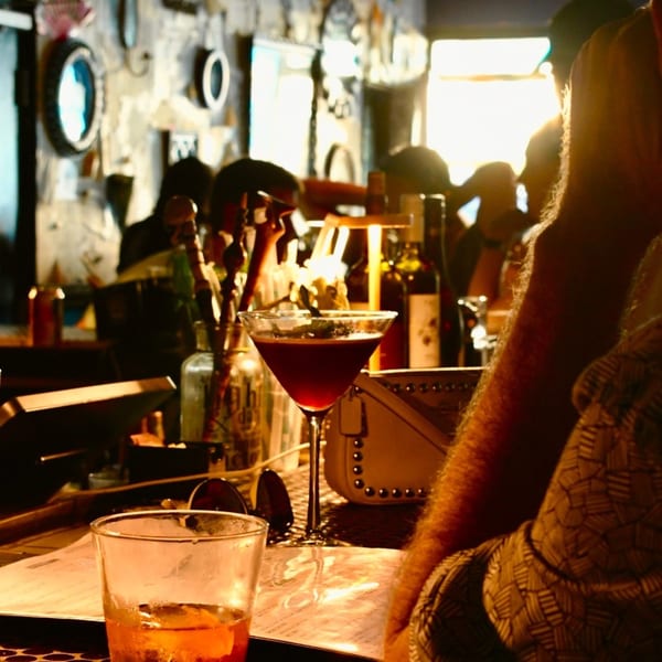 A red cocktail in a martini glass on a bar with patrons in the background.