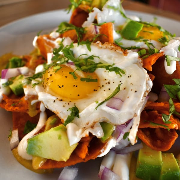 Chilaquiles with two fried eggs, avocado, red onion, sour cream, and cilantro on a white plate.