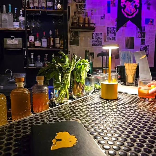 A dimly lit bar with bottles, fresh herbs, a cocktail, and a menu on a textured counter.
