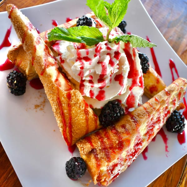 Fried dessert chimichangas with raspberry sauce, whipped cream, fresh mint, and blackberries on a white plate.