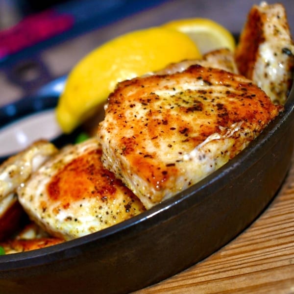 Close-up of seasoned chicken pieces with lemon wedges in a black skillet on a wooden surface.