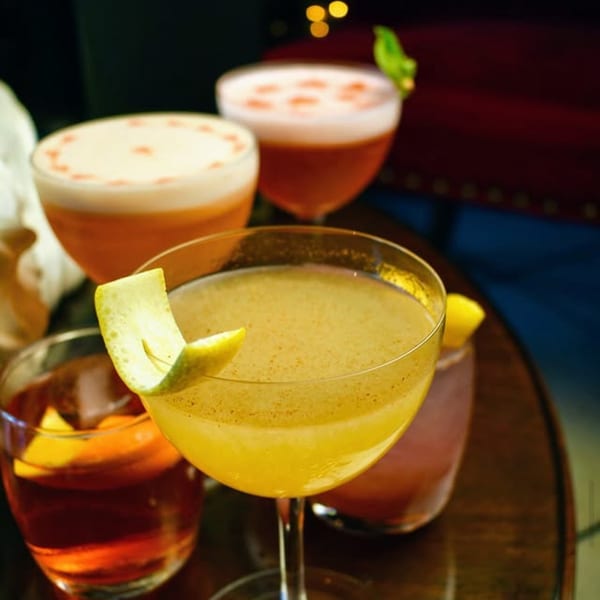 Four colorful cocktails, two with foamy tops and two with citrus garnishes, on a dark table.