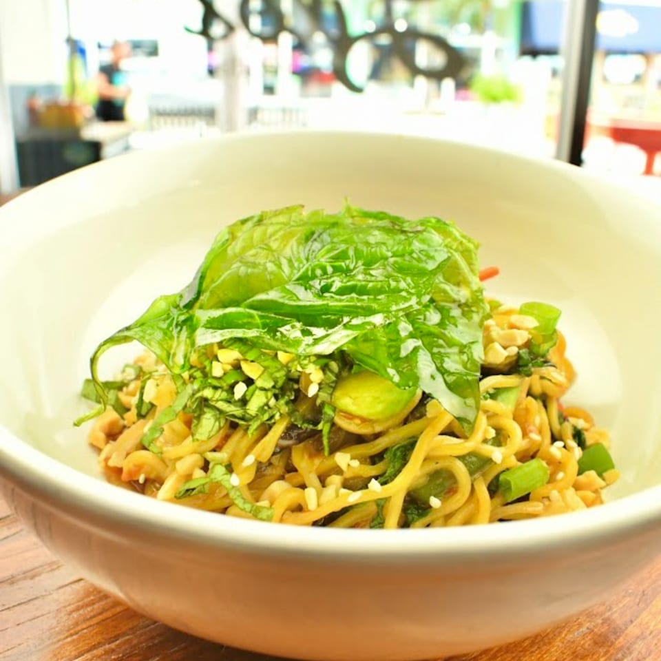 Noodle dish topped with fresh green basil, chopped nuts, and sesame seeds in a white bowl.