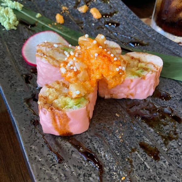 Pink sushi rolls topped with orange roe and white sprinkles, served on a dark plate with sauce.