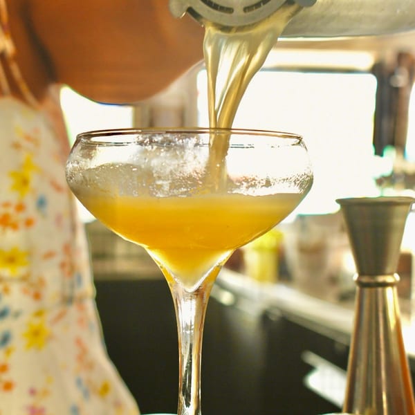 A bartender pours a vibrant yellow cocktail from a shaker into a coupe glass.