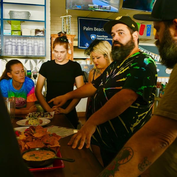 Customers and staff gathered around a bar counter with plates of nachos and other American dishes.