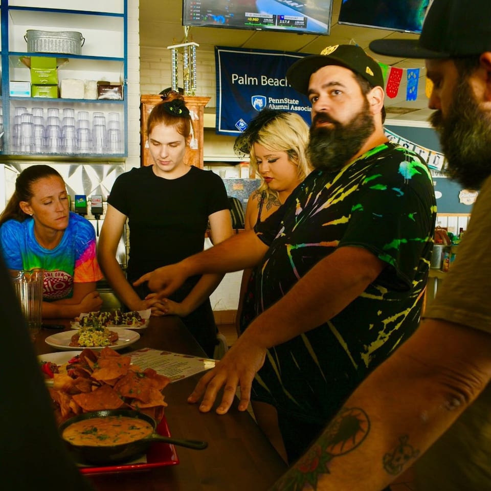 Customers and staff gathered around a bar counter with plates of nachos and other American dishes.