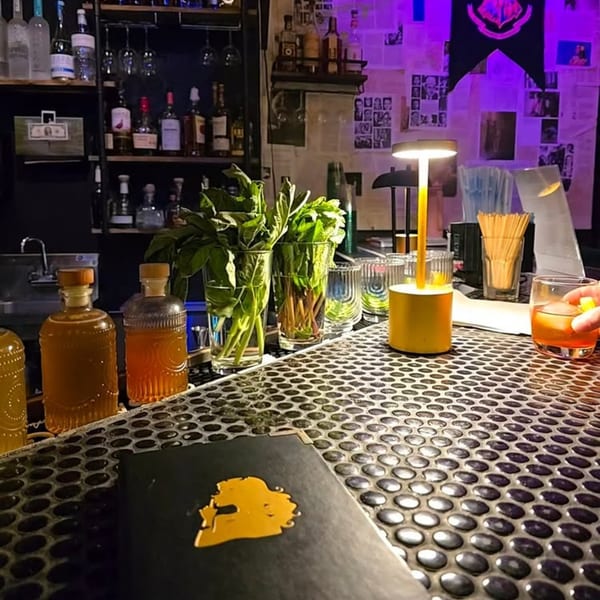 A dimly lit bar counter with bottles, fresh herbs, a cocktail, and a menu.