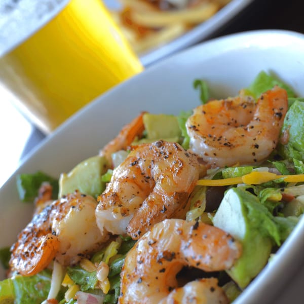Close-up of a shrimp and avocado salad with a blurred beer and fries in the background.