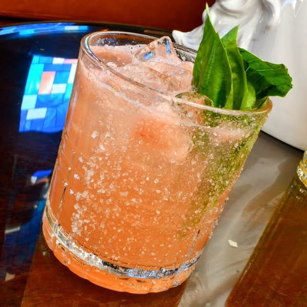 A vibrant orange cocktail with ice and basil garnish, served in a salt-rimmed glass.