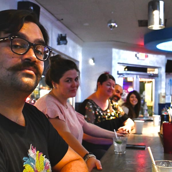 A group of friends, including a man with glasses and mustache, sitting at a bar counter.