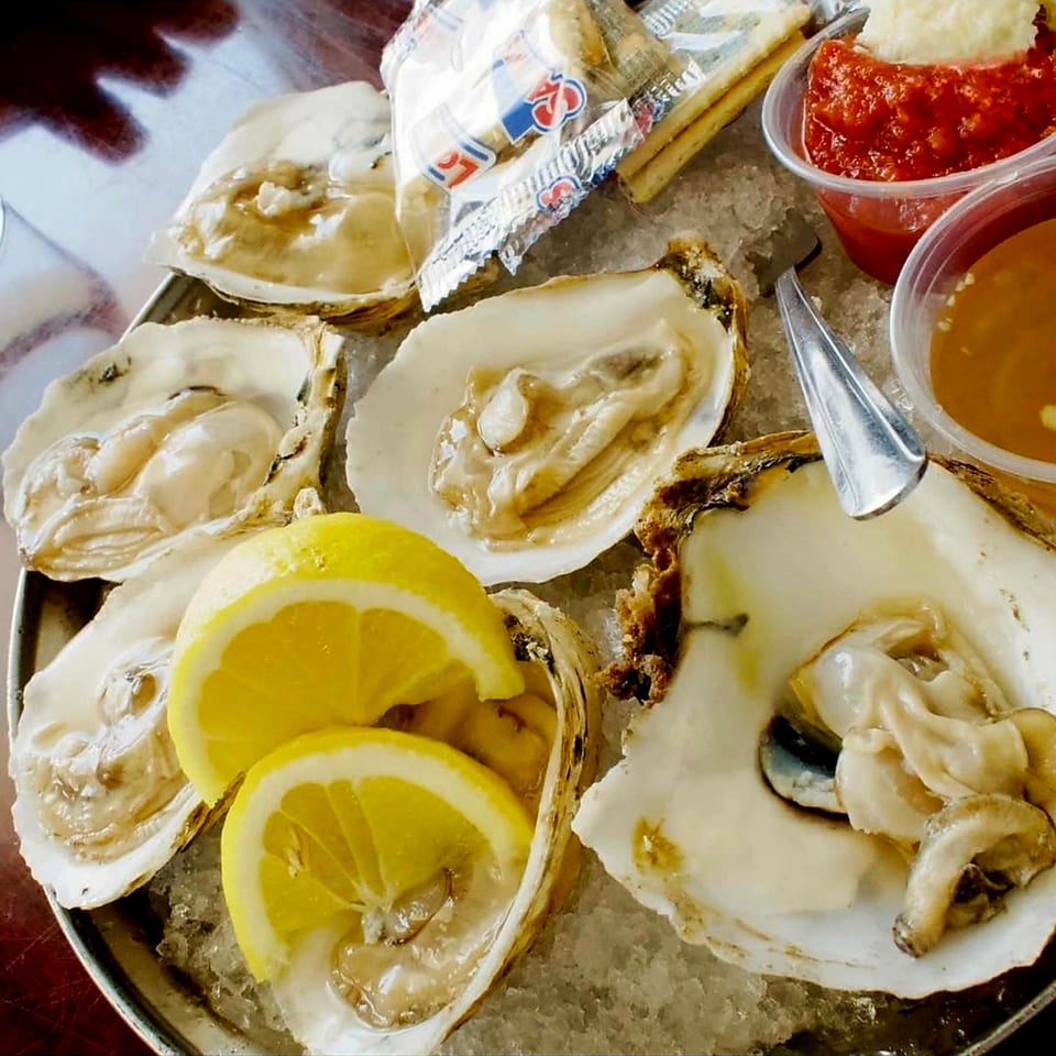 Fresh oysters on ice with lemon wedges, cocktail sauce, and crackers on a metal tray.