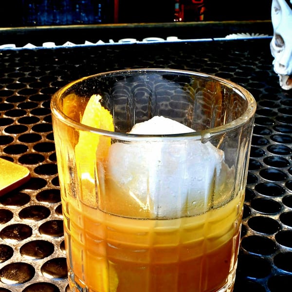 An Old Fashioned cocktail with a large ice cube and orange peel on a dark, textured bar.