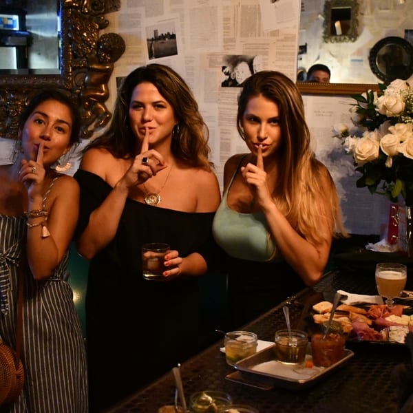Three women at Lilo's Streetfood & OKA Sushi pose with fingers to lips, surrounded by drinks and appetizers.